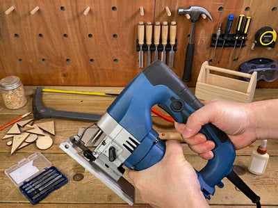 Woodworker using a blue electric jigsaw to cut lumber on a workbench in a DIY workshop