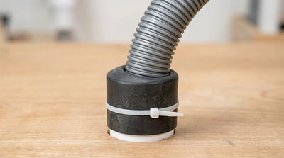 A close-up of a completed DIY drain pipe seal on a wooden floor, featuring a grey corrugated flexible hose inserted into a main pipe, wrapped tightly with a thick black rubber sleeve and secured by a white nylon zip tie | HomeDIYer